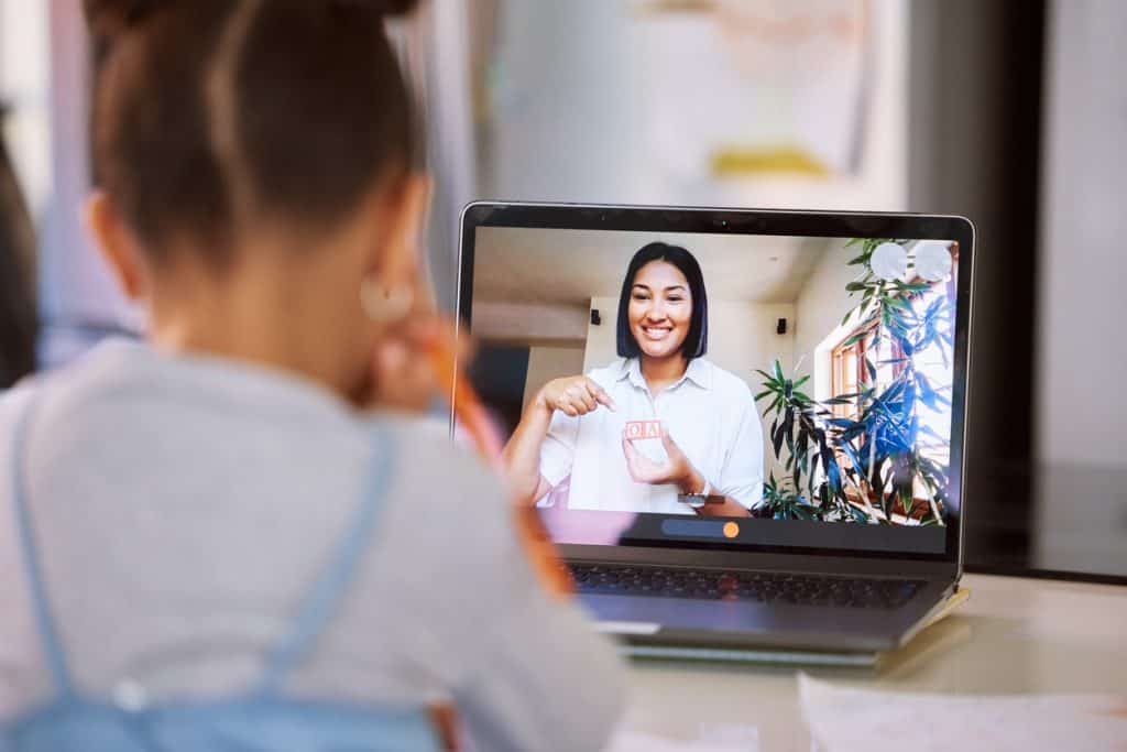 Little Girl On A Video Call With Her Teacher Using A Laptop. Young Female Teacher Doing A Virtual Lesson Using Blocks With Her Student. Little Girl Doing A Virtual Speech Therapy Session With A Woman
